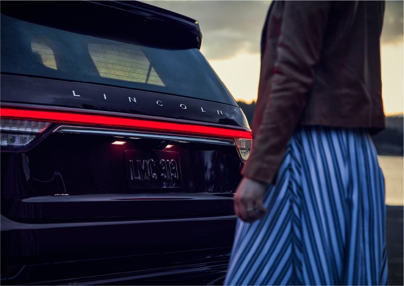 A person is shown near the rear of a 2023 Lincoln Aviator® SUV as the Lincoln Embrace illuminates the rear lights | Caruso Lincoln in Long Beach CA