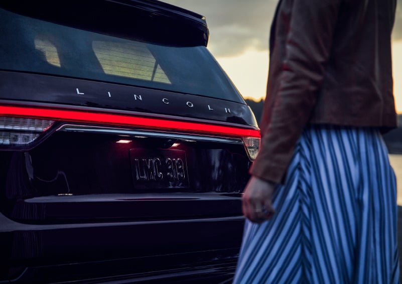 A person is shown near the rear of a 2024 Lincoln Aviator® SUV as the Lincoln Embrace illuminates the rear lights | Caruso Lincoln in Long Beach CA