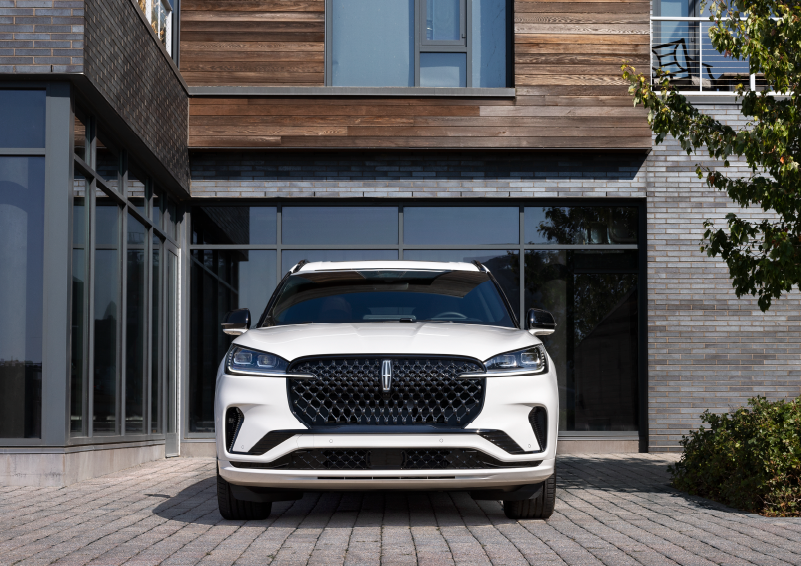 A white 2025 Lincoln Aviator® SUV is shown parked outside a modern building with gray brick and wood accents. | Caruso Lincoln in Long Beach CA
