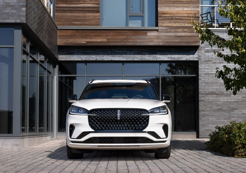 A white 2025 Lincoln Aviator® SUV is shown parked outside a modern building with gray brick and wood accents. | Caruso Lincoln in Long Beach CA