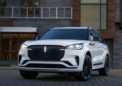 A white 2025 Lincoln Aviator® SUV is shown parked outside a modern building with gray brick and wood accents. | Caruso Lincoln in Long Beach CA