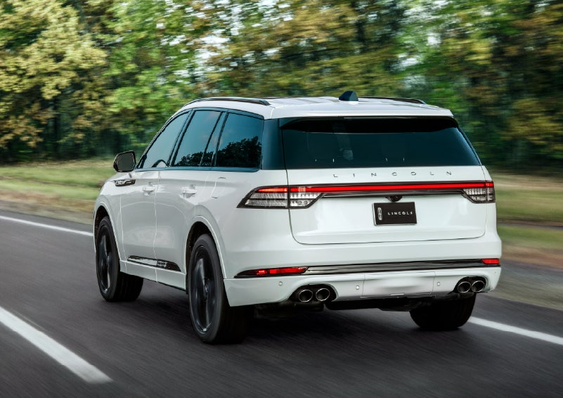 A white 2026 Lincoln Aviator® SUV is being driven away down a road lined with colorful trees. | Caruso Lincoln in Long Beach CA